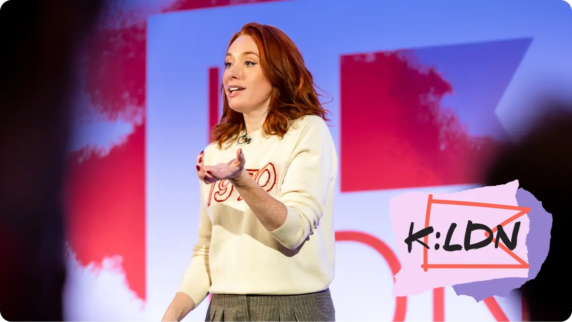 A person speaking on stage with a microphone, wearing a white sweater. The background features the text "K:LDN".
