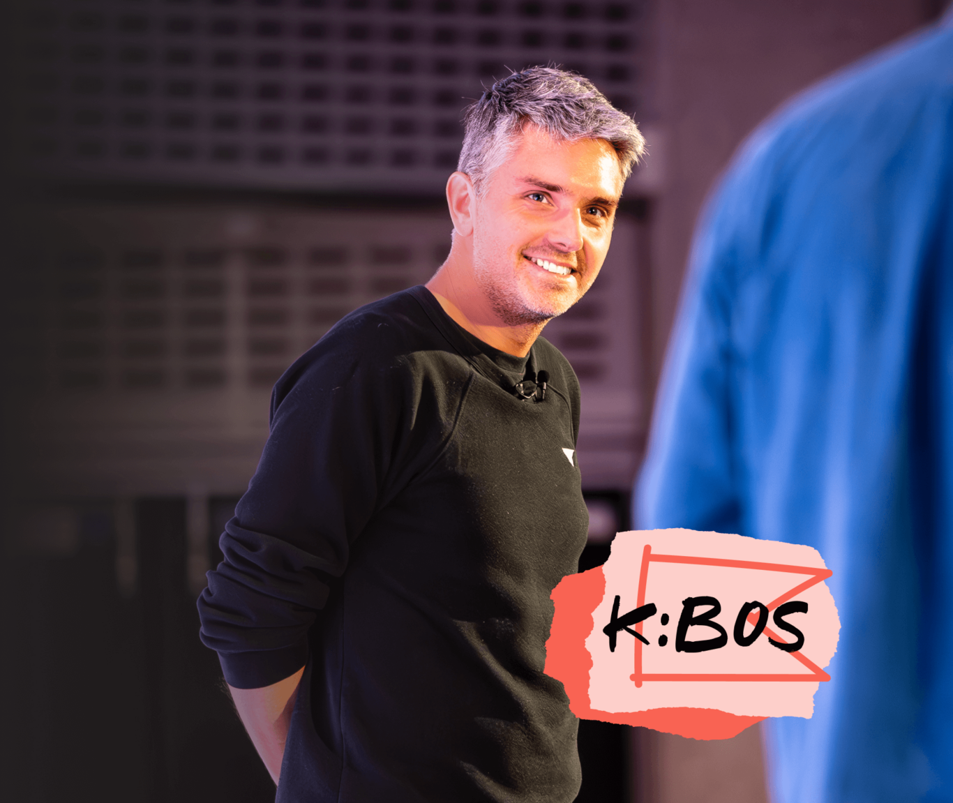 A smiling man in a black shirt stands on stage with a microphone. The text "K:BOS" is displayed in the corner.