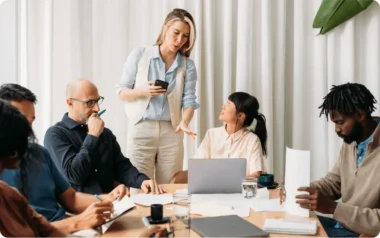 A group of people in a meeting, with one person standing and speaking, while others sit and take notes.
