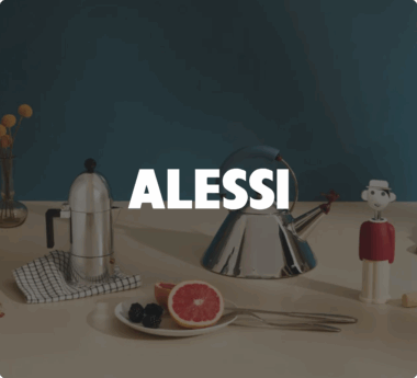 Stoviglie Alessi posizionate su un tavolo, tra cui un bollitore, una macchina per il caffè e un piatto da frutta. Il nome &quot;ALESSI&quot; è ben visibile.