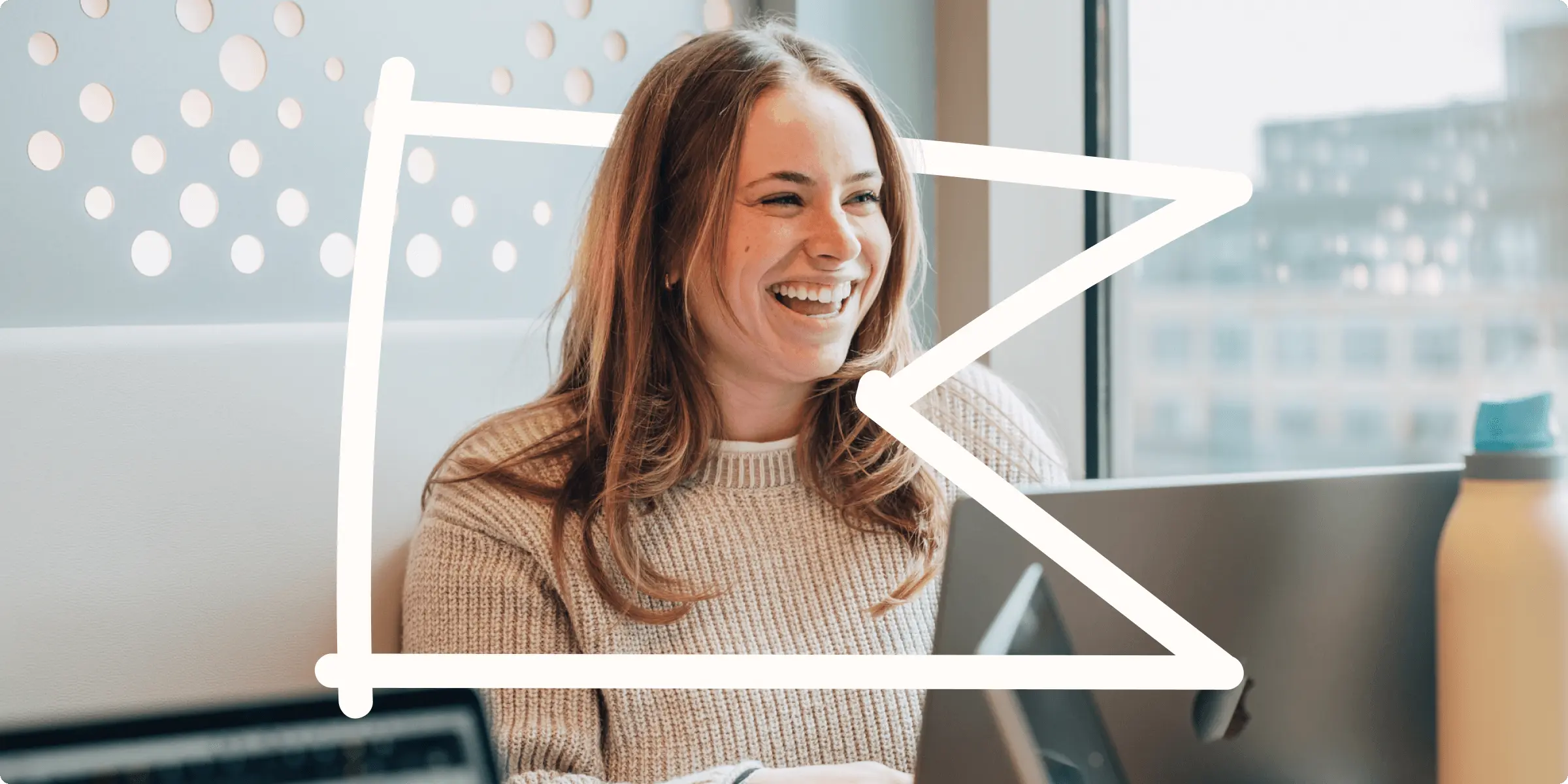 Smiling woman sitting at a desk with a laptop, wearing a sweater, next to a window.