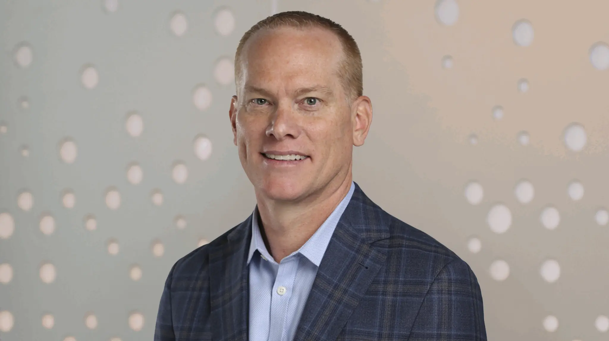 Smiling man in a blue plaid suit jacket and light blue shirt, standing against a patterned background.